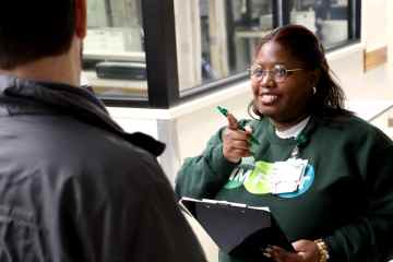 A woman in a dark green sweatshirt with "Sim First" written on the front holding a clipboard while talking to a man in a gray jacket