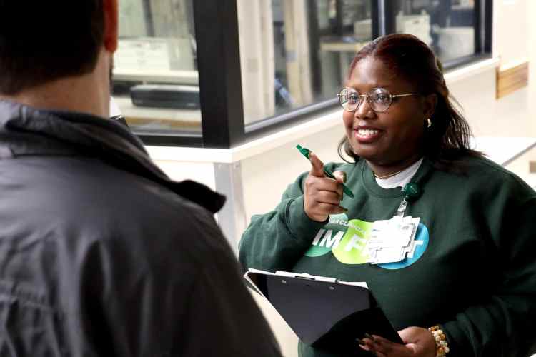 A woman in a dark green sweatshirt with "Sim First" written on the front holding a clipboard while talking to a man in a gray jacket
