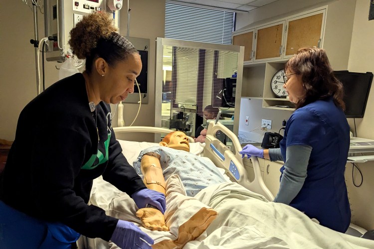 Two women in navy blue medical scrubs participating in a health-care simulation with a health-care simulation manikin