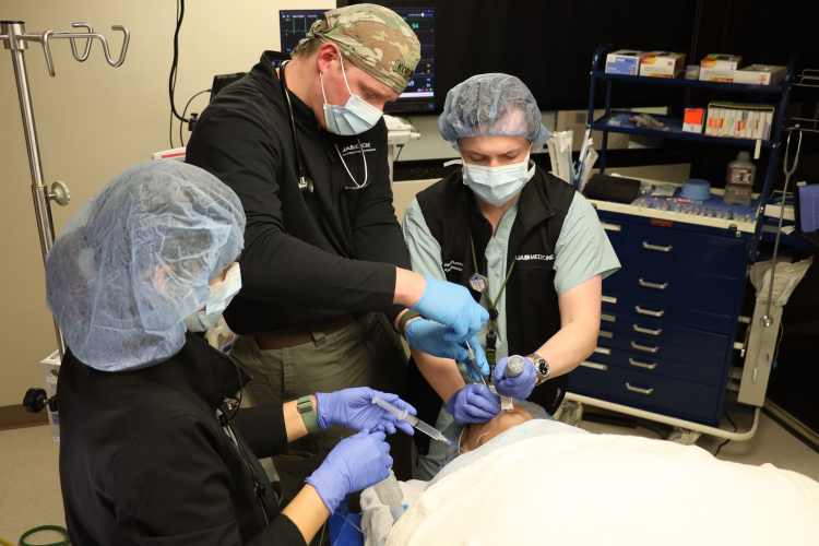 Three health-care professionals—two female and one male—participating in a health-care simulation with a health-care simulation manikin in a simulated operating room