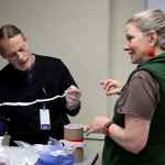 A man in black medical scrubs participating in a procedural coaching simulation and a woman in a gray T-shirt and green vest coaching him