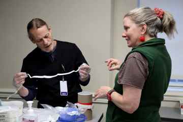 A man in black medical scrubs participating in a procedural coaching simulation and a woman in a gray T-shirt and green vest coaching him