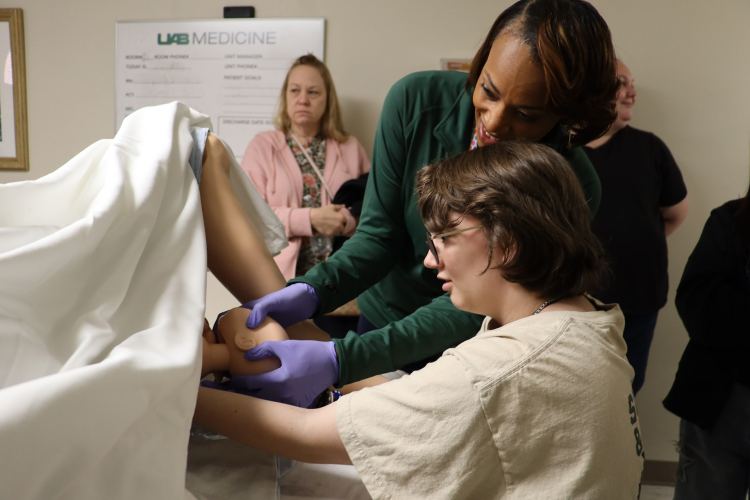 A female OB professional in a green jacket assists a Girl Scout in a cream-colored T-shirt participate in a simulated childbirth