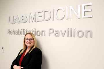 A woman in a business suit standing in front of a wall with lettering that reads "UAB Medicine Rehabilitation Pavilion."