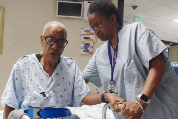 An elderly woman in a hospital gown receiving assistance walking by a female nurse in light blue medical scrubs