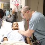 A woman in gray medical scrubs and a man in light blue medical scrubs interacting with a health-care simulation manikin laying in a hospital bed
