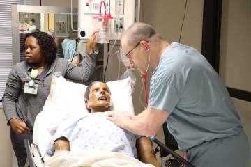 A woman in gray medical scrubs and a man in light blue medical scrubs interacting with a health-care simulation manikin laying in a hospital bed
