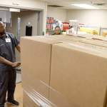 A man in gray medical scrubs pushing a pallet containing stacked cardboard boxes into a supply room