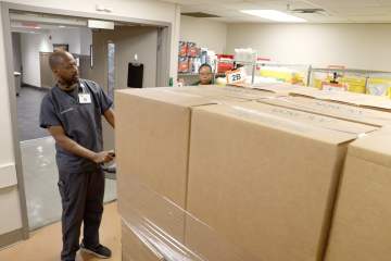 A man in gray medical scrubs pushing a pallet containing stacked cardboard boxes into a supply room