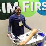 A nurse in navy blue scrubs, standing behind a wheeled cart holding healthcare simulation equipment