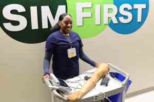 A nurse in navy blue scrubs, standing behind a wheeled cart holding healthcare simulation equipment