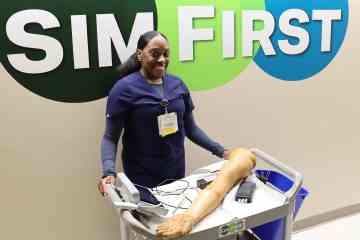 A nurse in navy blue scrubs, standing behind a wheeled cart holding healthcare simulation equipment