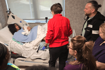 A female standardized patient portraying a new mother holding a newborn baby manikin, laying in a hospital bed and interacting with various members of the health-care team