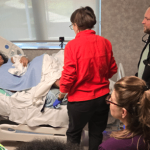 A female standardized patient portraying a new mother holding a newborn baby manikin, laying in a hospital bed and interacting with various members of the health-care team