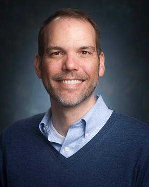 A headshot of a caucasian male wearing a light blue collared shirt and a navy blue v-neck sweater