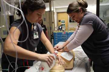 Two female music therapists in gray medical scrubs performing CPR on a healthcare manikin, as another music therapist in gray medical scrubs observes in the background