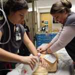 Two female music therapists in gray medical scrubs performing CPR on a healthcare manikin, as another music therapist in gray medical scrubs observes in the background