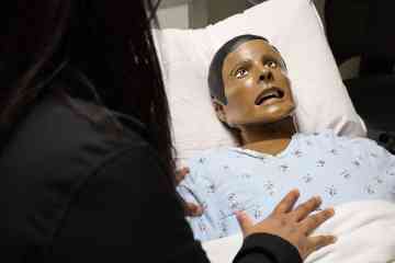 A health-care professional seen from behind, addressing a healthcare manikin in a hospital bed during a simulation