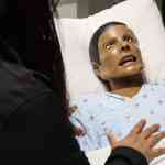 A health-care professional seen from behind, addressing a healthcare manikin in a hospital bed during a simulation