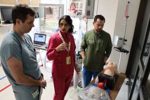 A man in light blue medical scrubs, a woman in fuchsia medical scrubs and a man in a blue medical scrubs and a green jacket standing together, assessing a healthcare simulation trainer.
