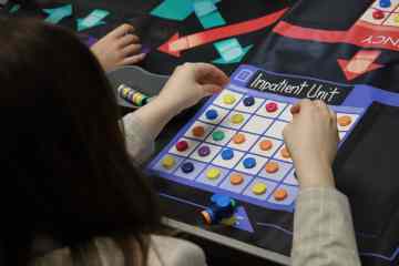 Pictured from behind, a woman with dark hair placing tiles on a gameboard that reads "Inpatient Unit," as part of a healthcare simulation.