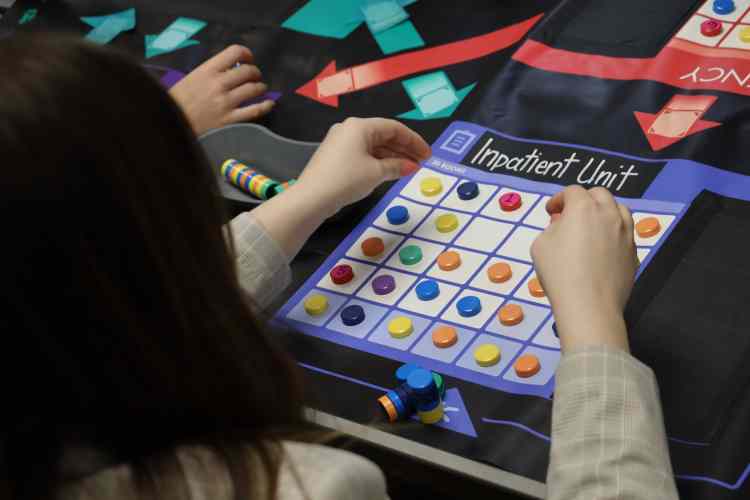 Pictured from behind, a woman with dark hair placing tiles on a gameboard that reads "Inpatient Unit," as part of a healthcare simulation.