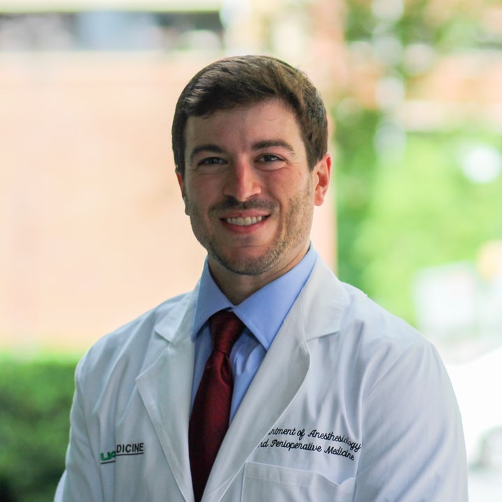A Caucasian man with brown hair wearing a white physician's lab coat, a blue shirt and red tie.