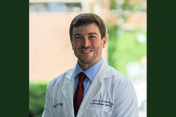 A Caucasian man with brown hair wearing a white physician's lab coat, a blue shirt and red tie.