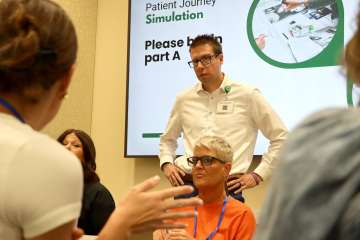 A male facilitator in a white shirt stands among seated participants engaged in a group discussion during a healthcare systems simulation exercise.