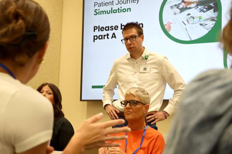A male facilitator in a white shirt stands among seated participants engaged in a group discussion during a healthcare systems simulation exercise.
