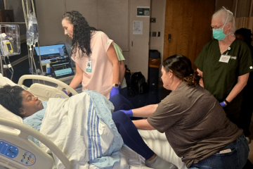Three healthcare professionals participate in a bedside simulation exercise with a standardized patient in a hospital room setting.