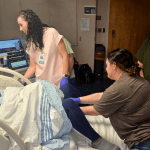 Three healthcare professionals participate in a bedside simulation exercise with a standardized patient in a hospital room setting.