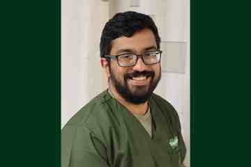 A dark-haired, bearded man wearing olive green medical scrubs
