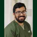 A dark-haired, bearded man wearing olive green medical scrubs