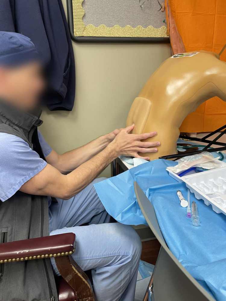 A male healthcare professional wearing light blue scrubs and a gray vest participating in a procedural healthcare simulation with a lumbar puncture training device