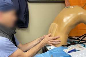 A male healthcare professional wearing light blue scrubs and a gray vest participating in a procedural healthcare simulation with a lumbar puncture training device