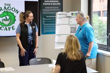 A woman in blue scrubs and a black UAB Medicine vest speaks with a female standardized patient in a blue T-shirt as another participant sits nearby.