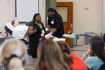 A healthcare educator presents to participants during a simulation workshop. A patient mannequin rests in a hospital bed nearby while another instructor demonstrates a clinical procedure at the front of the room.