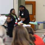 A healthcare educator presents to participants during a simulation workshop. A patient mannequin rests in a hospital bed nearby while another instructor demonstrates a clinical procedure at the front of the room.
