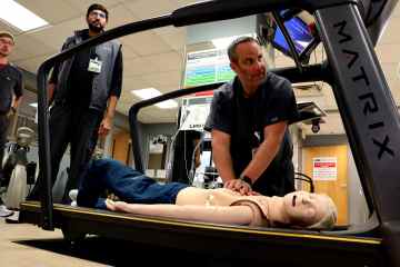 Healthcare professional performs chest compressions on a healthcare manikin positioned on a treadmill while other team members observe.