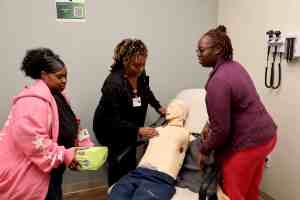 Three participants practice an emergency response scenario with a healthcare manikin, including preparing an AED for use.
