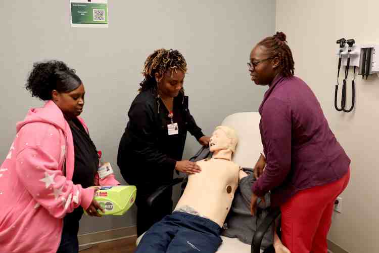 Three participants practice an emergency response scenario with a healthcare manikin, including preparing an AED for use.