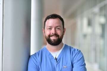 Portrait of a person in blue scrubs standing in a brightly lit hallway.