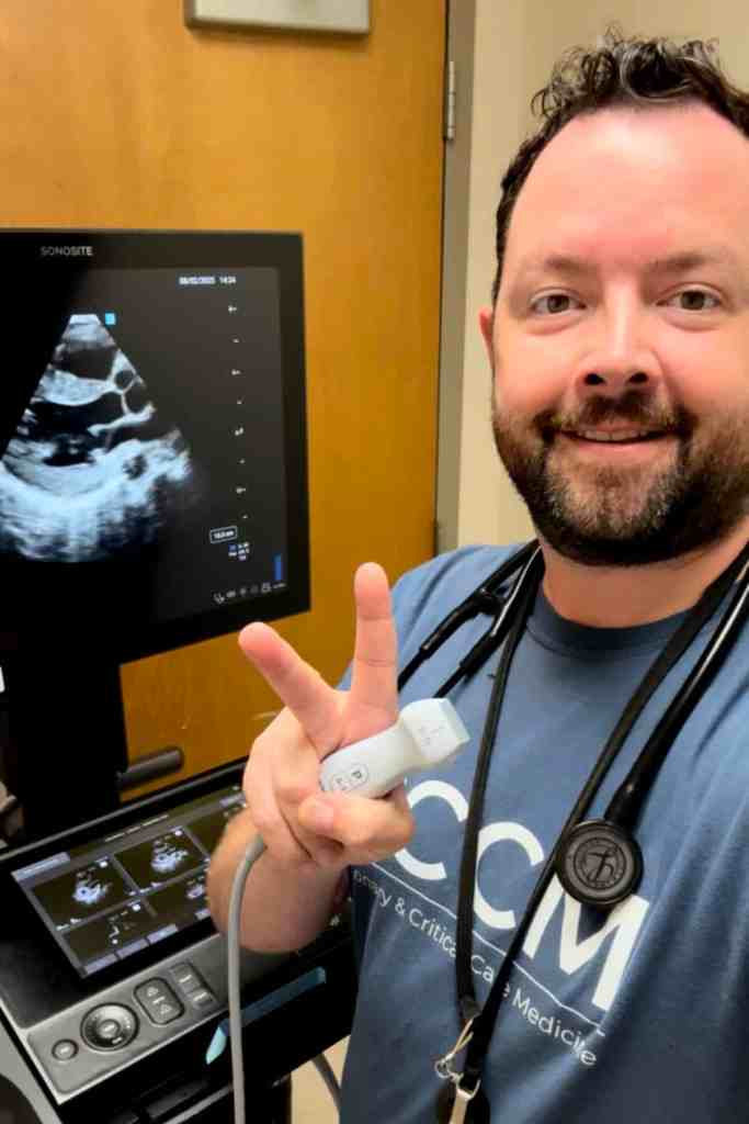 Person wearing a stethoscope holds an ultrasound probe beside an ultrasound machine displaying a cardiac ultrasound image.