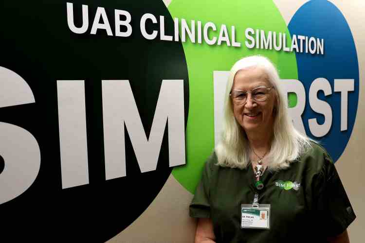 Woman in green scrubs standing in front of a colorful UAB Clinical Simulation Sim First wall display.