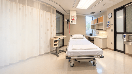 Clinical simulation room with a healthcare bed, privacy curtain, bedside equipment, wall-mounted medical equipment, cabinets, sink and computer workstation.