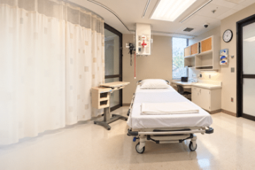 Clinical simulation room with a healthcare bed, privacy curtain, bedside equipment, wall-mounted medical equipment, cabinets, sink and computer workstation.